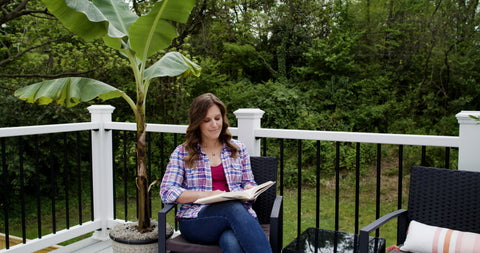 A woman reading under a banana tree in a cozy deck seating area.