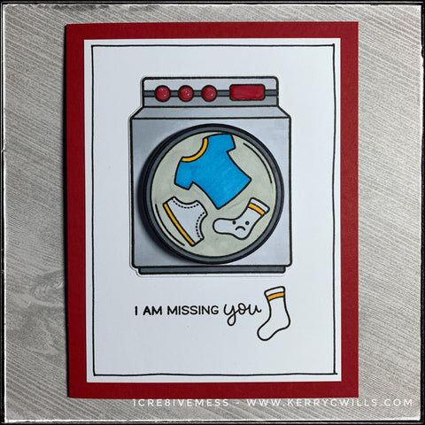 A flat-lay view of this handmade card designed to be sent to a friend or loved one that's being missed. A washing machine that has been stamped and die-cut is the center focal point. The middle drum is elevated off of the card base and wiggles back and forth. The sentiment "I am missing you" is stamped next to a lonely little sock. Hand drawn lines surround the perimeter of the white card front on top of the red card base. 