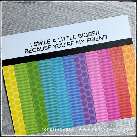 An angled view of the card front - all the colors are visible in their vertical stripes and polka dotted or striped patterns. The sentiment is stamped in black ink which is very bold along the white open space. It reads "I smile a little bigger because you're my friend." and is sure to make the recipient smile. What a great way to share some happiness. 