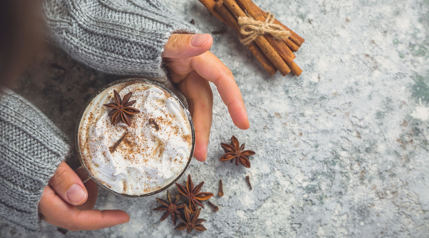 Collombatti Naturals 8 reasons to love chai this winter blog - arms wearing a grey woollen jumper going to cup a mug of chai on a grey background with a bundle of cinnamon sticks and star anise in front of the cup