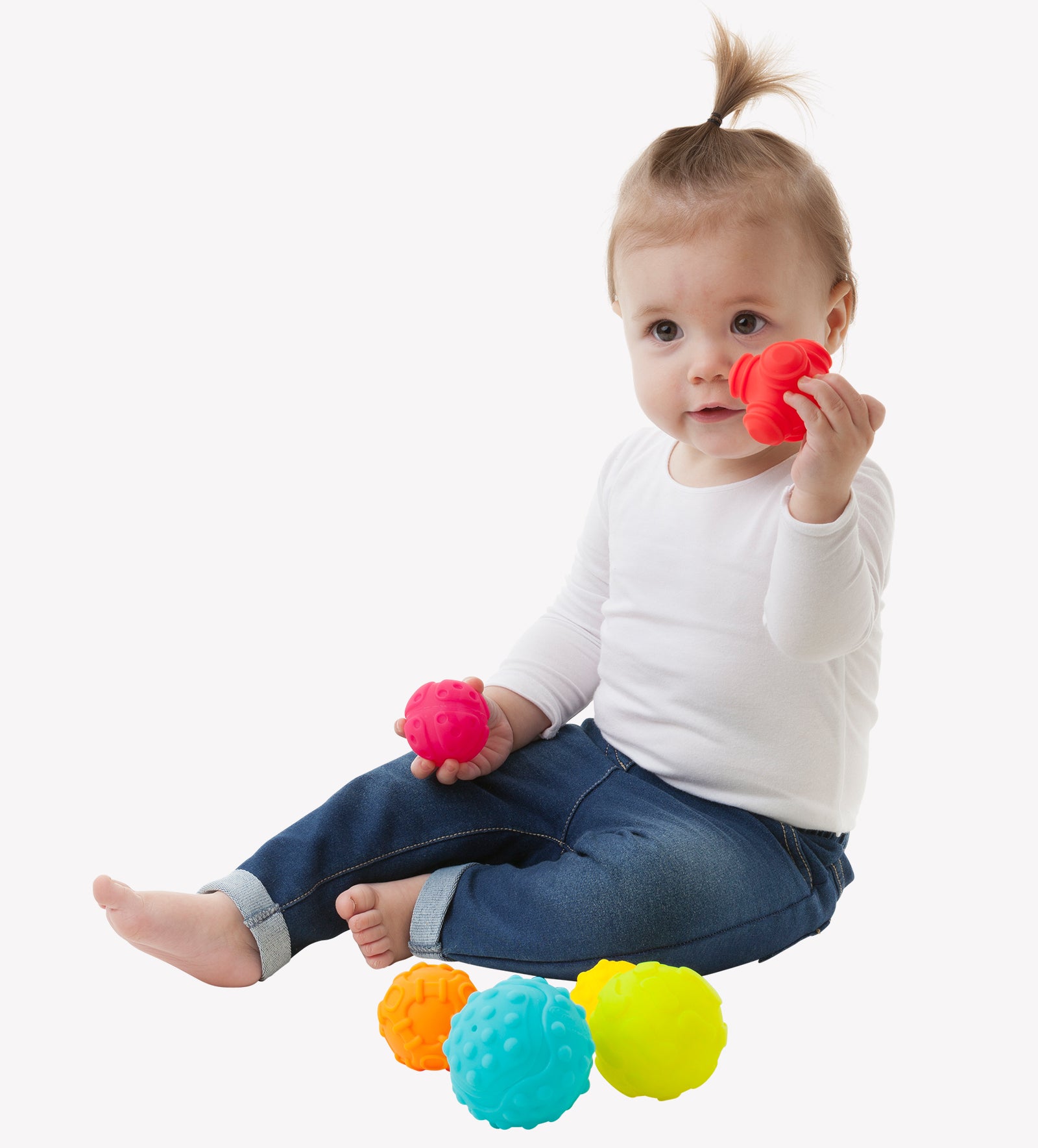 A toddler girl sits down on the floor playing colorful Playgro Textured Ball while holding the red ball