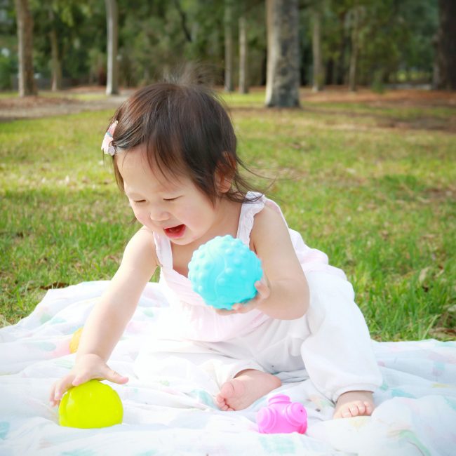 A toddler girl is playing with Playgro textured ball toys outdoor on the garden