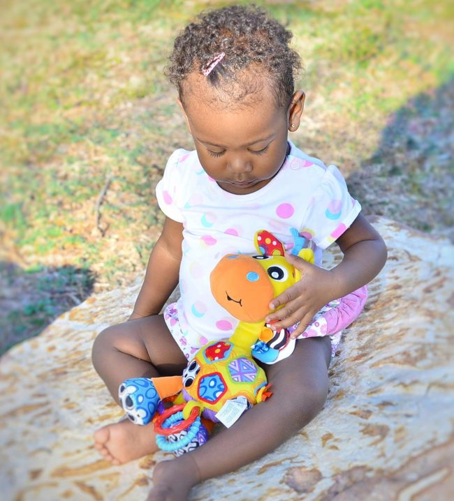 A toddler is sitting outdoor on the rocks holding Playgro Giraffe baby toys in her lap bought from Not Too Big Online Store