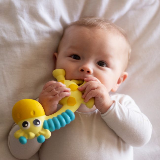 A baby lying down in bed chewing on Playgro Jerry baby teether