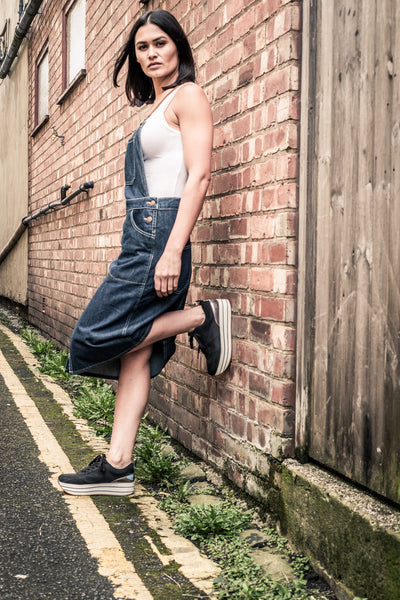 Leaning against red brick wall wearing denim bib overall dress.