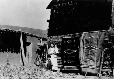 Photo of author's great grandmother with author's father standing in front of handmade woven blankets and chile ristras in Chimayo, circa 1936