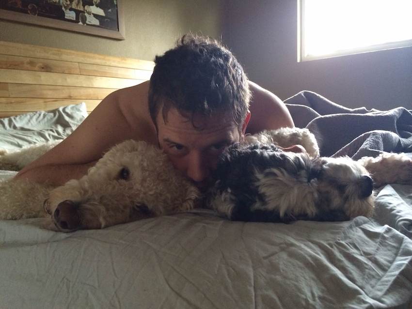 A handsome man and two dogs snuggle together on a bed.