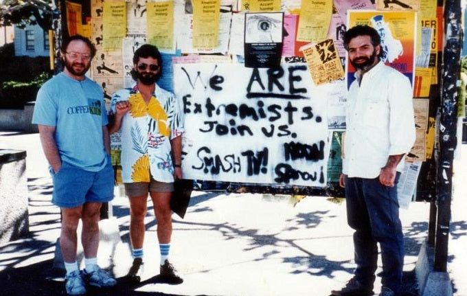 Image: young Dean wearing a Hawaiian shirt, standing in front of a bulletin board with two male friends. A sign on the bulletin board says "We ARE extremists. Join us."