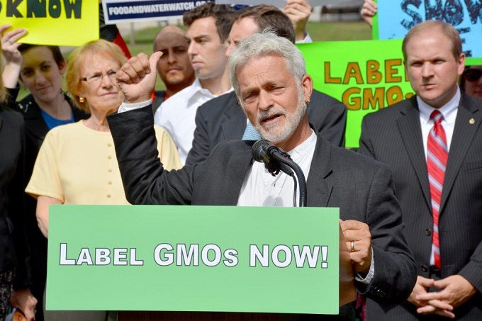 Image: Dean standing at a podium in front of a crowd holding signs. A sign on the front of the podium reads "Label GMOs NOW!"