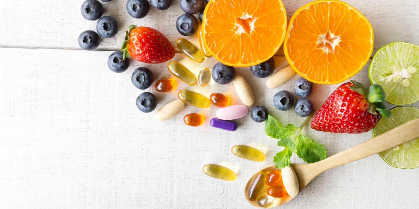 A wooden spoon holding a variety of multivitamin pills among a backdrop of fruits