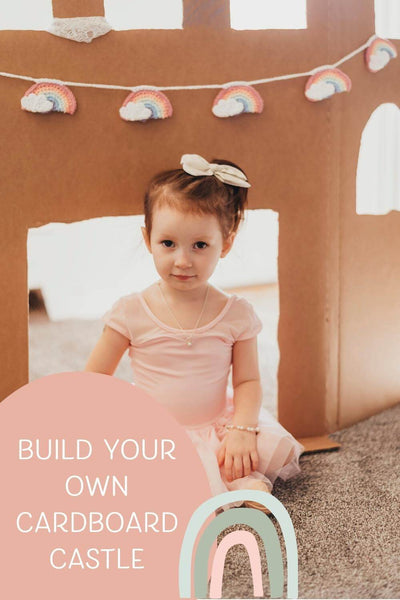 little girl wearing jewelry from little girl's pearls sitting in front of a cardboard castle with a rainbow garland.