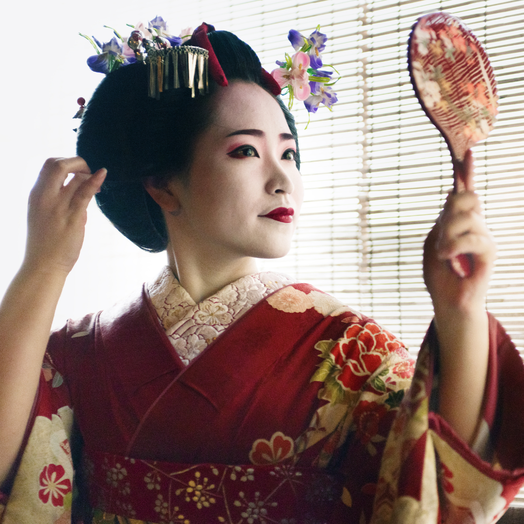 Eine Geisha mit opulentem Haarschmuck schaut in einen Handspiegel