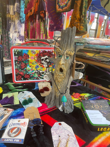 A small sculpture of a tree with a face with handmade necklaces and bracelets wrapped around it surrounded by other goods (handing tie dye, incense holders, socks, lunch tins, etc) under an artists tent