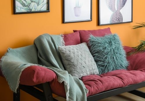 Wall painted orange with a modern burgundy couch against the wall, and throw pillows and blankets of grey and teal. These colours are spicy, and full of flavor.
