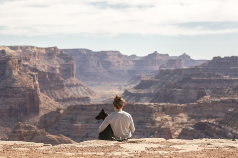 Woman sits on cliff overlooking canyons with an arm around his adopted dog.