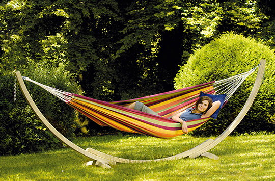 A woman sat on her bright red, orange, yellow and light green hammock on a wooden stand, in her garden.