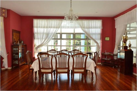 A dining room with red painted walls, white curtains and a light brown dining room table and chairs.