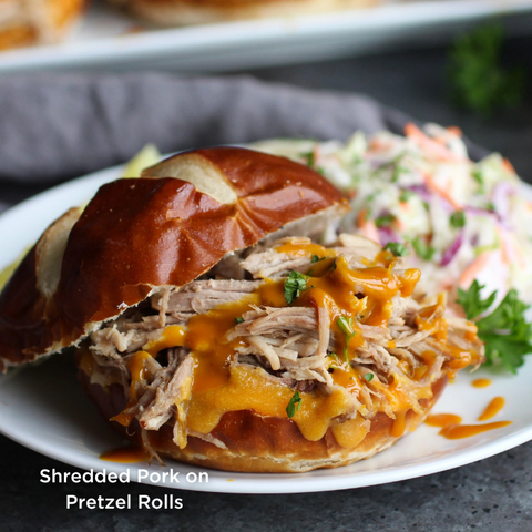 Shredded Pork on Pretzel Rolls. Image depicts Shredded Pork with melted shredded cheese in between pretzel buns. Served on a white plate with a side of coleslaw.