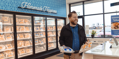 Image shows a man walking through the Let's Dish! store with the freezer cases and "Homemade Happiness" sign in the background. Man is carrying two meals and smiling while looking away from the camera.