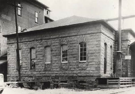 A black and white photo of the Dalles Mint. A one story building with 5 windows on 1 side. 
