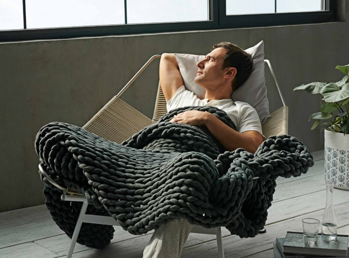 Man lounging on a chair underneath a Bearaby Napper weighted blanket