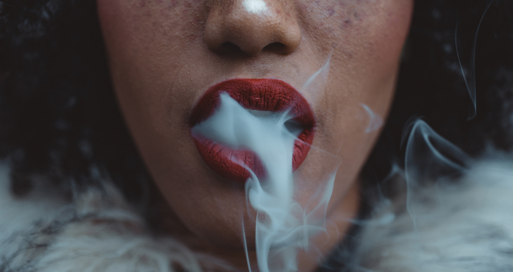 woman with red lipstick, dark brown curly hair, and freckles exhaling a cloud of smoke from a close up angle