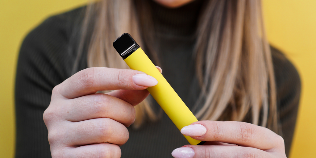 A woman with a dark green sweatshirt holding a yellow disposable vape in the foreground in focus with her body and yellow background slightly out of focus