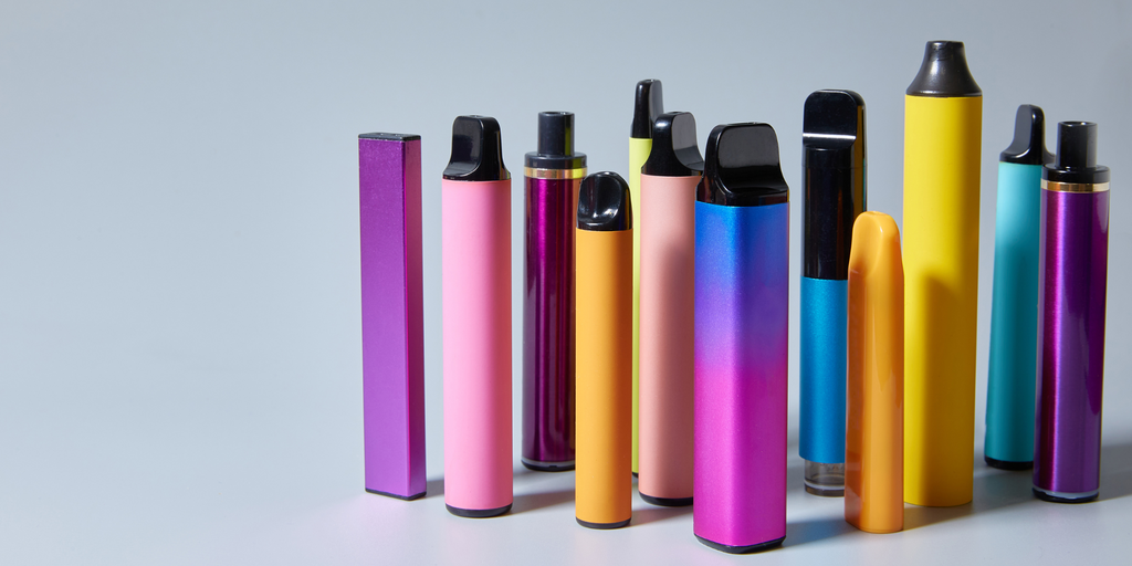 A bunch of multi colored disposable vapes standing upright in a group with a blank background and surface