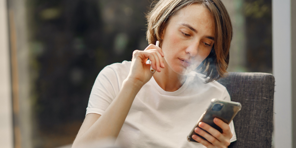 A woman in a white t-shirt looking at a phone in one hand while holding a disposable vape in the other exhaling smoke in a rural background