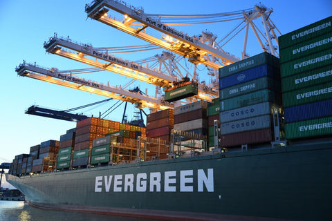 Cargo shipped with the name 'Evergreen' painted onto the hull with hundreds of cargo shipping containers stack on the bow with cranes hovering above with writing 'Port of Baltimore'