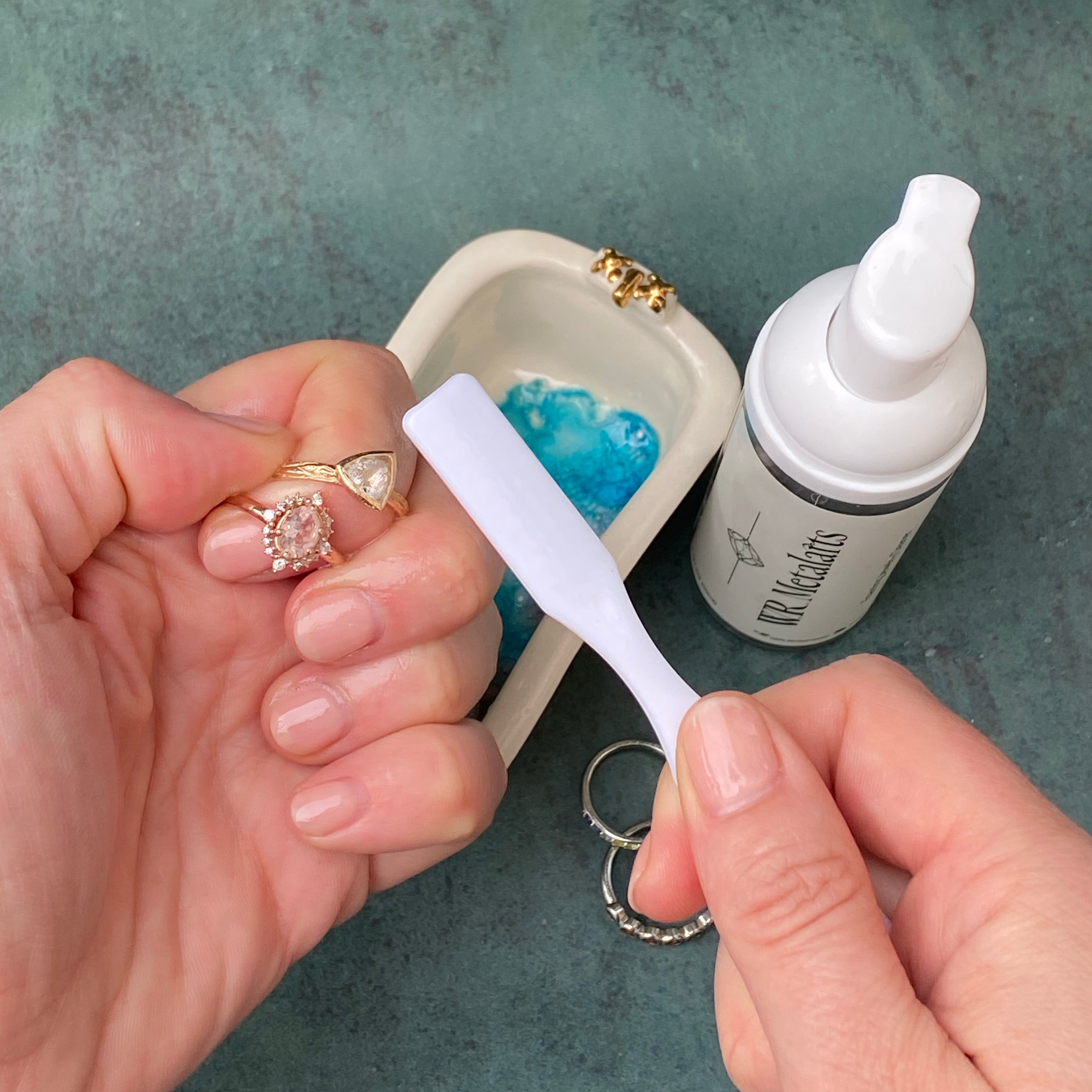 Hands using soft white brush to clean several rings with W.R. Metalarts Jewelry Cleaner