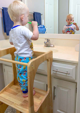 Child standing on a learning toddler tower brushing teeth in the bathroom
