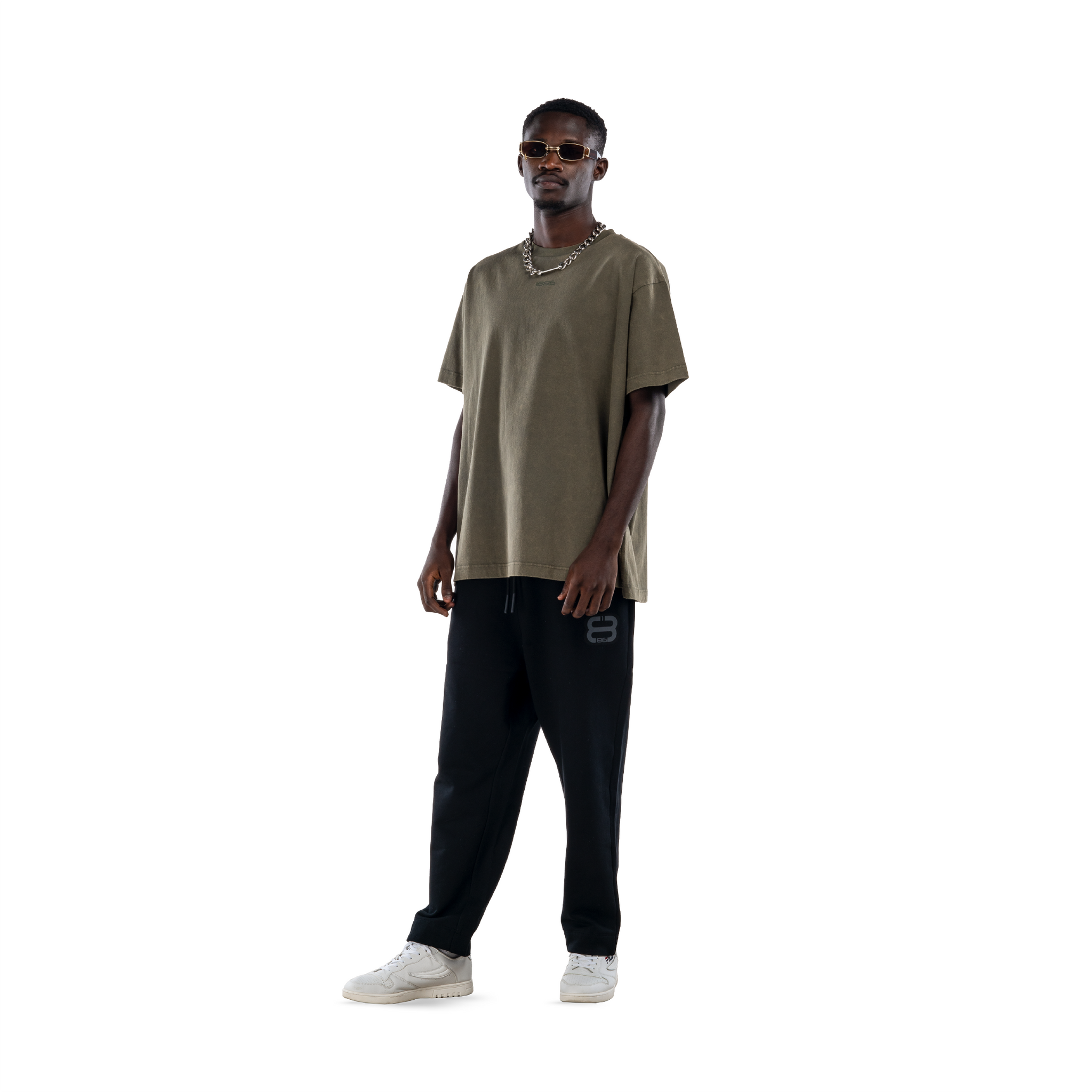 model wears olive green t-shirt with black sweatpants, sunglasses and white shoes. front view.