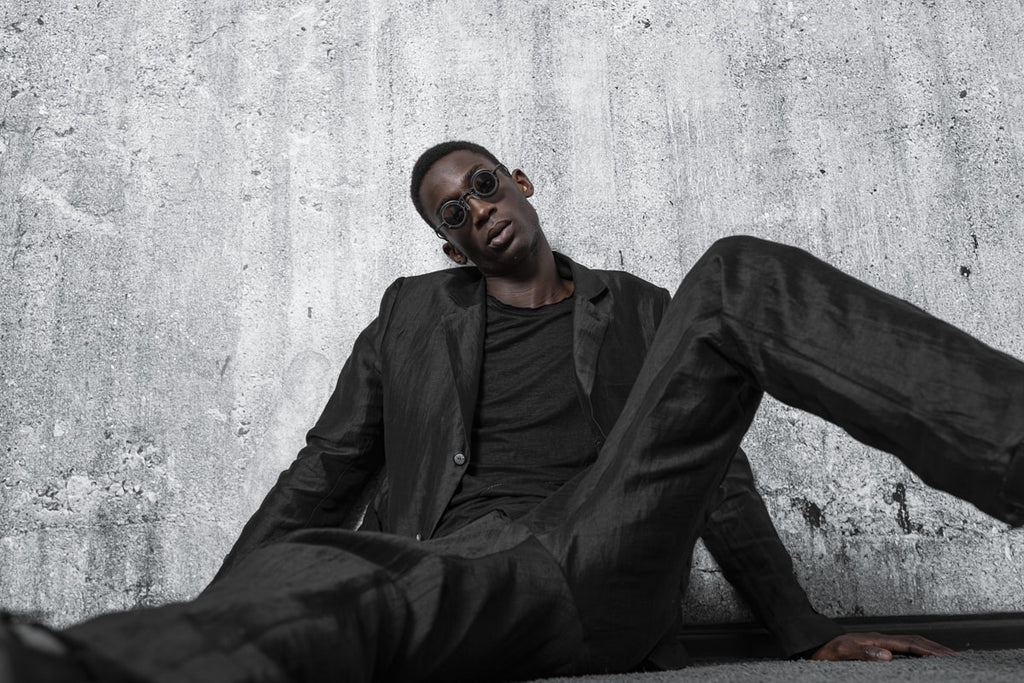 Ein Mann sitzt am Boden mit Designer Sonnenbrille in einen All Black Outfit im Dark Avantgarde Mode Stil von eigensinnig wien