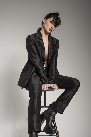 Eine Frau sitzt auf einem Stuhl vor einem weißen Hintergrund mit einem All-Black-Hosenanzug von eigensinnig wien im Dark Avantgarde Stil