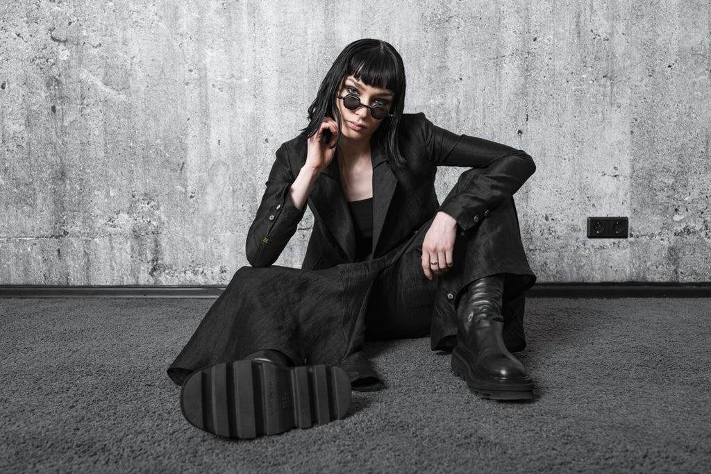 woman sits on the floor with designer sunglasses in an all black outfit in the dark avant-garde fashion style of eigensinnig wien