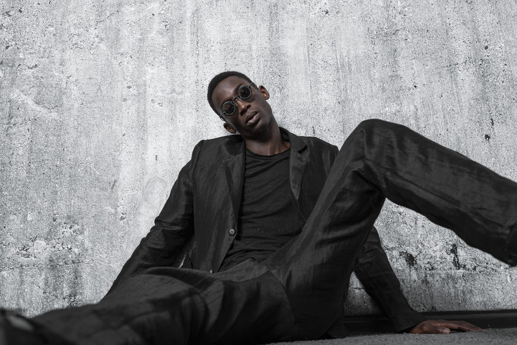 A man sits on the floor with designer sunglasses in an all black outfit in the dark avant-garde fashion style of eigensinnig wien.