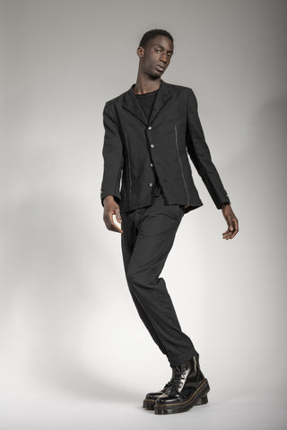 A man stands in front of a white background wearing an all-black suit by eigensinnig wien in dark avant-garde style