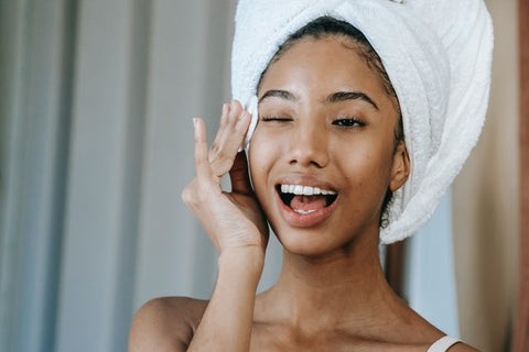 POC taking eye makeup off with a beautiful smile.  She has her hair wrapped up in a towel and looks as if she has just come from the shower.