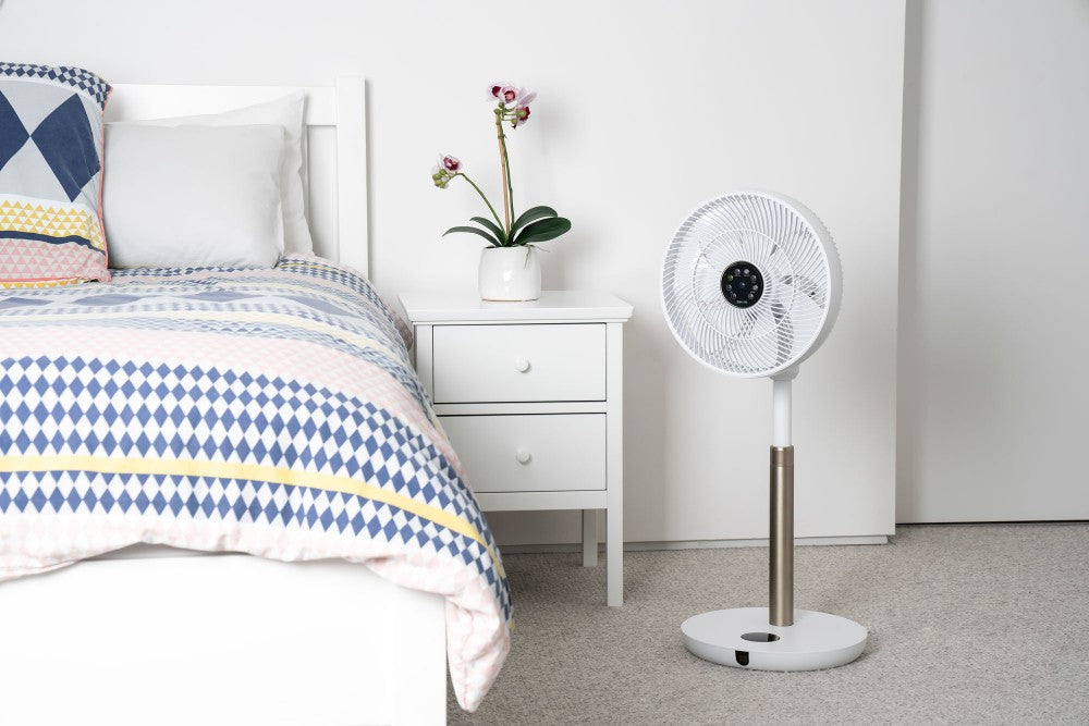 Lifestyle image of the Devola 12 inch fan next to a bed. There is a side table with an orchid. 