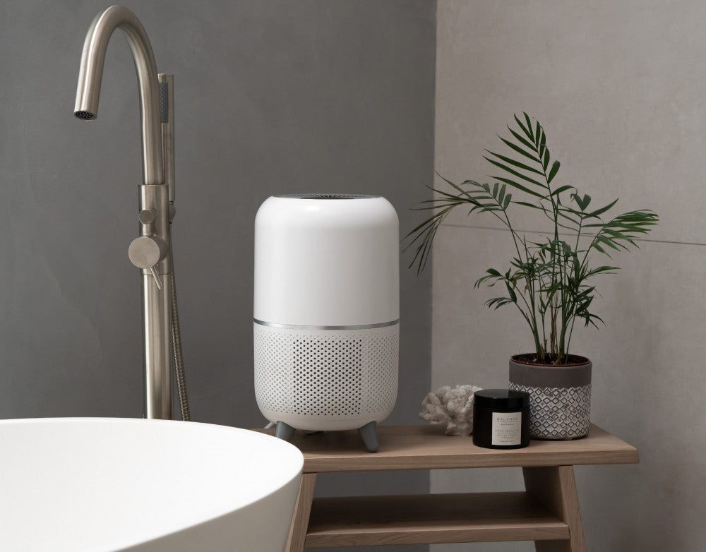 Lifestyle image showing the Devola air purifier with feet on a wooden sideboard in a bathroom. It is next to a white sink with a golden chrome tap.