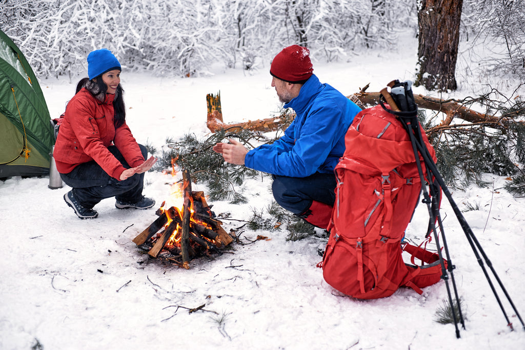 man and woman camping in the snow warming their hands by a fire