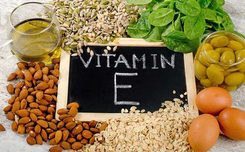 It is a square image. In the center there is a white board on which it says: "vitamin E". Around there are different foods rich in this vitamin such as eggs and almonds.