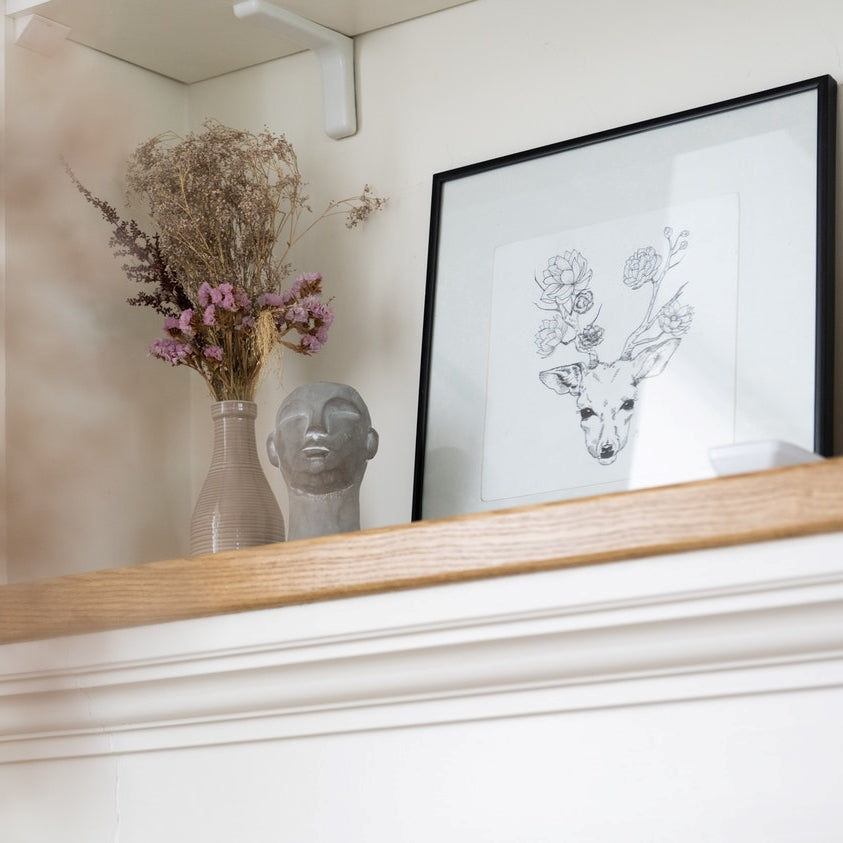 bouquet of dried flowers in a ceramic vase atop a mantelpiece beside a grey sculpture of a head and an artwork of a deer head with flower antlers
