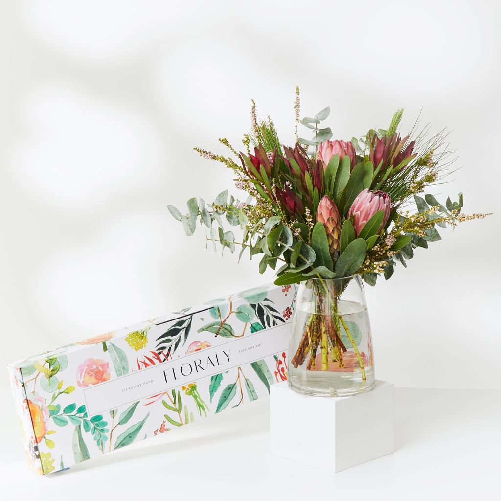 seasonal natives bouquet of protea, conebush, eucalyptus and other native flowers and foliage in a glass vase atop a white plinth beside a Floraly branded gift box