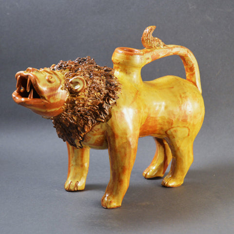 A ceramic vessel formed into a standing lion with its mouth open to reveal four vicious fangs.  The object is a water jug, the mouth is the spout, and the tail forms a handle by curving over to attach to the back of the lion.  The vessel is glazed in a honey-yellow colour.