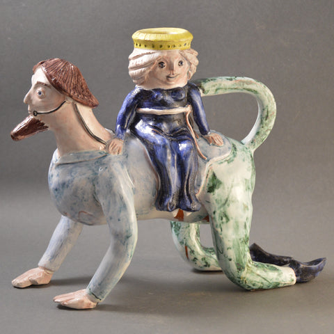 A ceramic vessel formed into a man crawling with a woman sitting on his back.   The man has a long, straight beard that forms the spout of this water jug.  The smiling woman is wearing a deep cobalt blue dress and a yellow-gold crown, while the man is in a pale blue tunic and green trousers.
