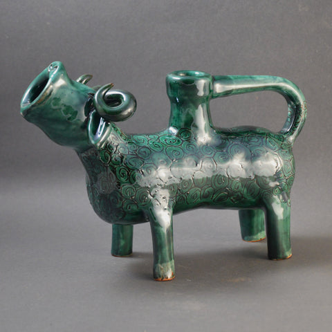 A ceramic vessel formed into a standing ram with its head turned up and its mouth open as if it is bleating.  The object is a water jug, the mouth is the spout, and the tail has been elongated to form a handle that curves around to attach to the back of the vessel.  The vessel is glazed in a lustrous green.
