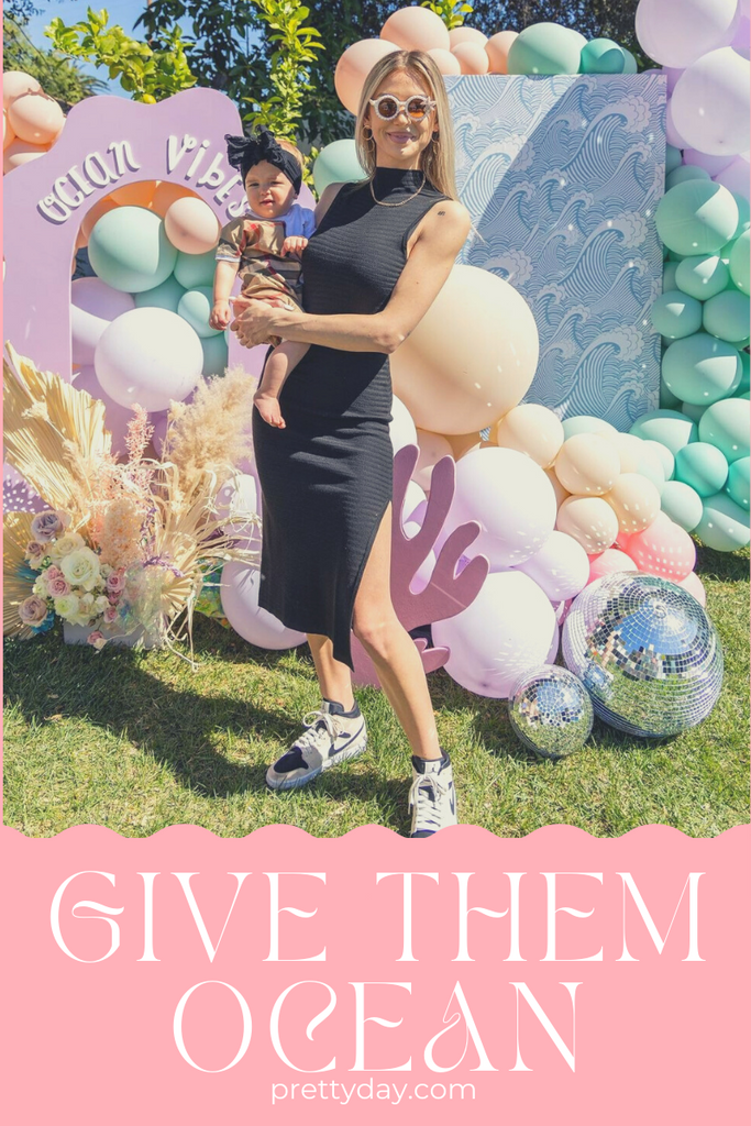 Give them ocean, Lala Kent, Mermaid themed party.  A woman in a black dress stands holding a toddler in front of a pastel balloon backdrop.