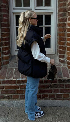 A woman standing sideways wearing medium wash denim, a white long sleeve shirt and a black vest